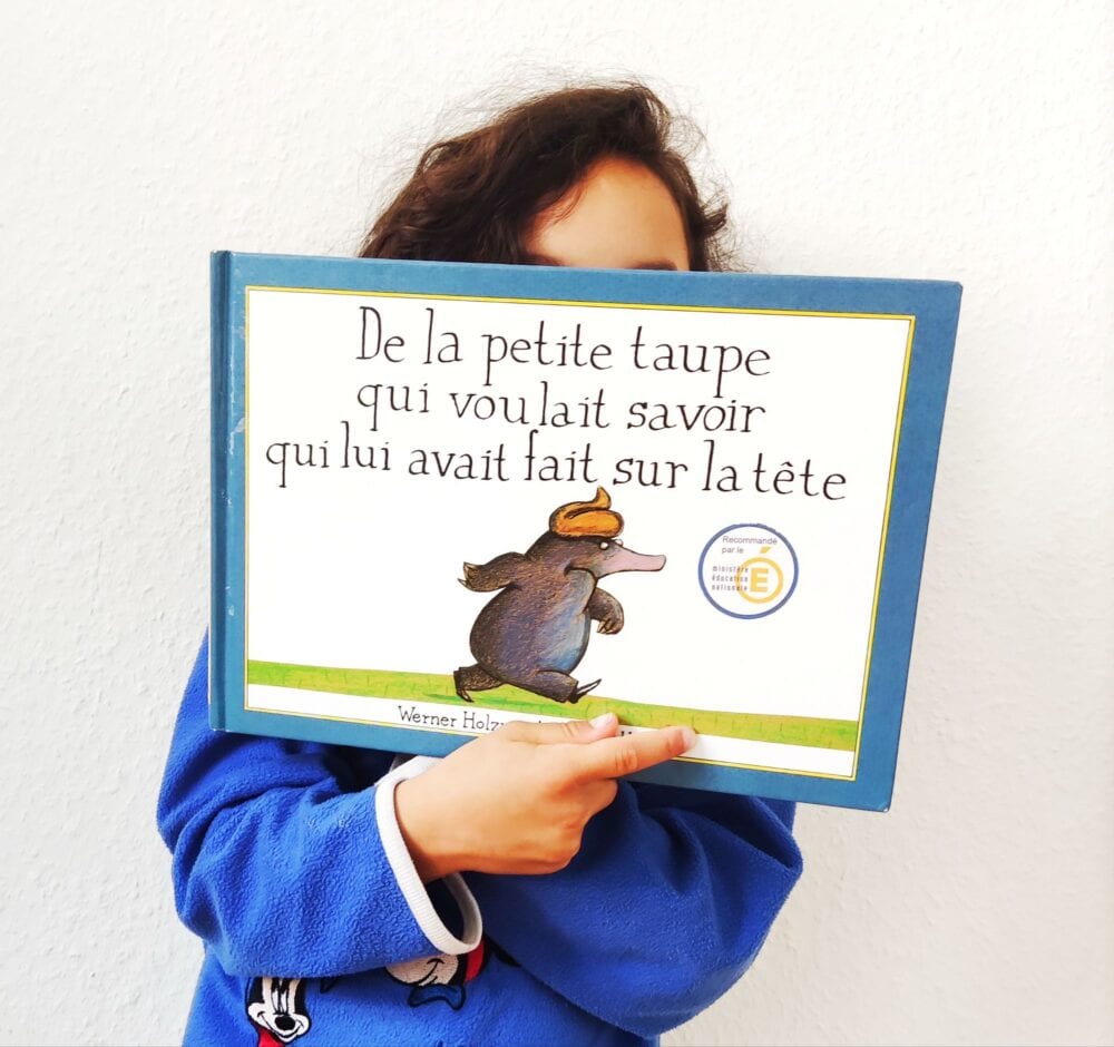 Couverture de l'album De la petite taupe qui voulait savoir qui lui avait fait sur la tête, illustrée par Wolf Erlbruch. On y voit une petite taupe noire, debout sur un monticule de terre, arborant une expression déterminée. Une crotte marron repose sur sa tête. Le fond est blanc, et le titre du livre est inscrit en haut de l'image.